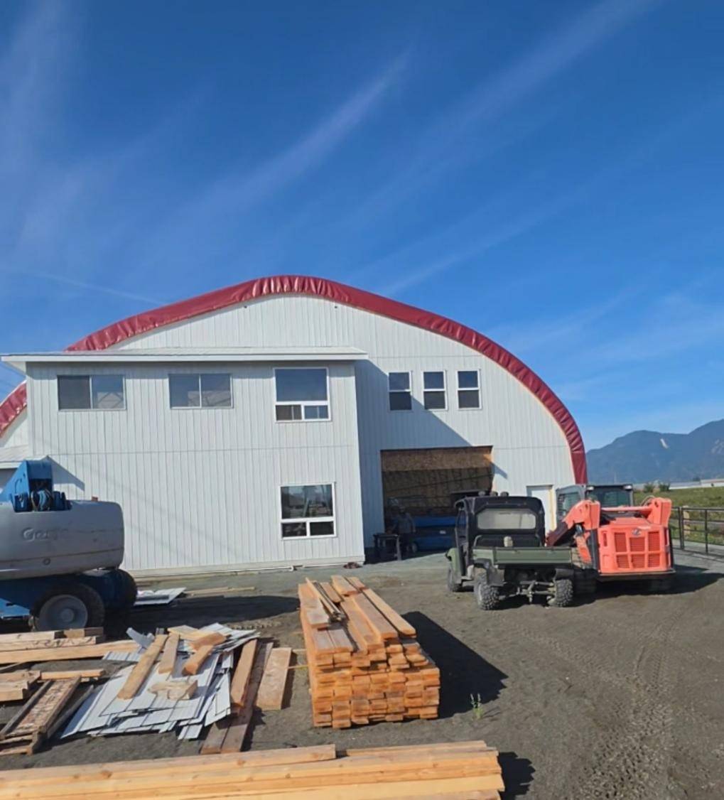 Arena shell in white metal under the red arch, lumber and machines in the yard