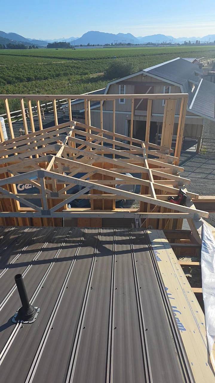 Roof trusses going up over a metal roof, with a barn and fields behind